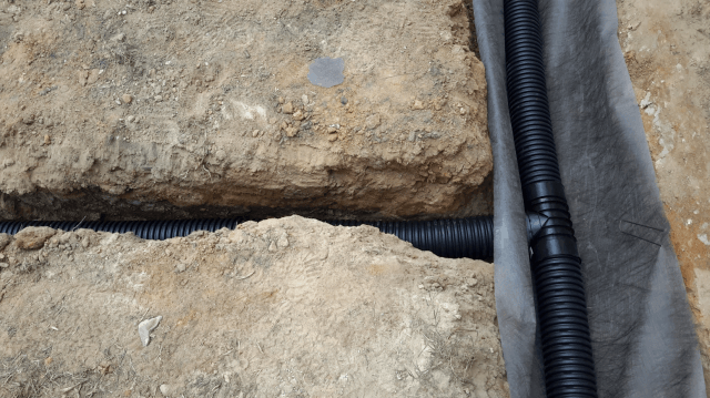 Black corrugated drain pipe in a trench, partially wrapped in grey fabric. Brown soil surrounds it.