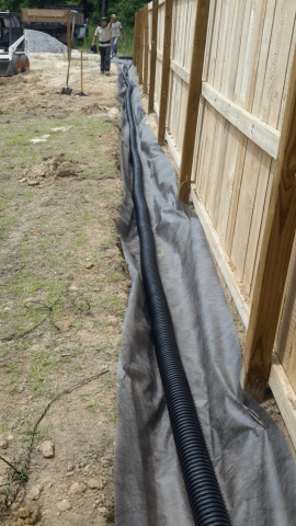 A black corrugated drainage pipe along a fence, wrapped in gray fabric. A person walks in the background.