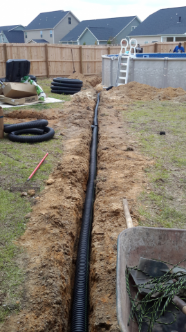 A trench in a backyard contains black drainage pipe, near a pool and fence. Construction tools and materials are visible.
