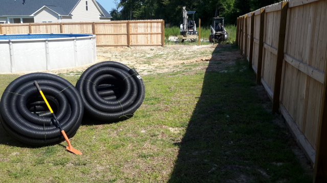 Backyard with above-ground pool, coiled black pipes, two excavators, and a wooden fence.
