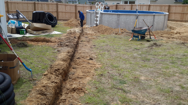 Person digging a trench in backyard with a pool. Dirt is piled up along the trench.
