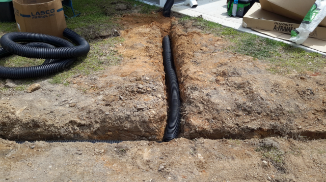 Trench with black corrugated pipe for drainage, outdoors on dirt and grass.