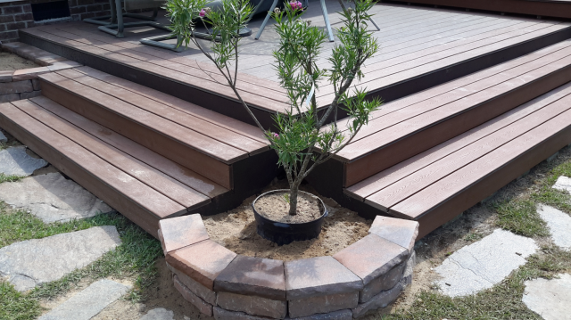 Wooden deck with brick-edged planter holding a small tree. Steps lead up to the deck, set in a grassy yard.