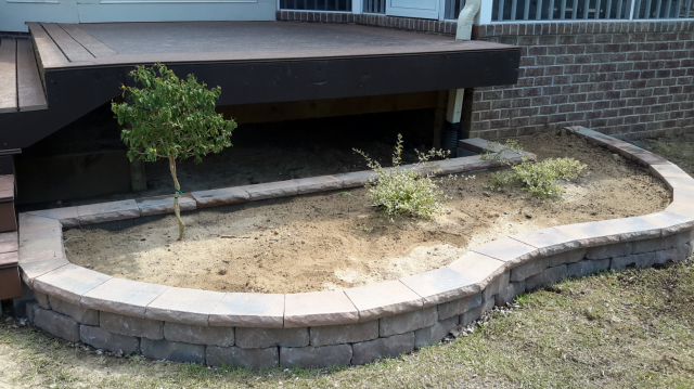 Curved brick retaining wall surrounds a bare garden bed under a wooden deck. A small tree and shrubs are planted.