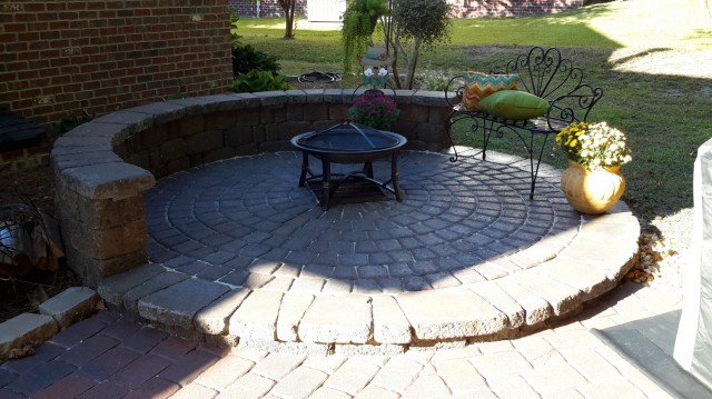 Circular brick patio with a fire pit, seating, and a low brick wall in a yard.