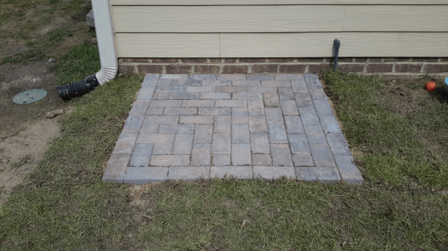 Square brick patio next to a house with grass surrounding it.