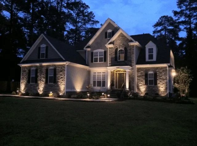 Lit-up house at dusk with stone facade and black shutters. Landscape lighting illuminates the front.