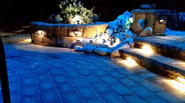Stone patio with illuminated retaining wall, steps, and water feature at night.