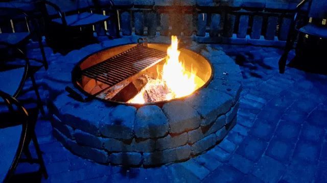 Fire pit with grill, flames burning brightly. Blue-toned patio with chairs in the background.