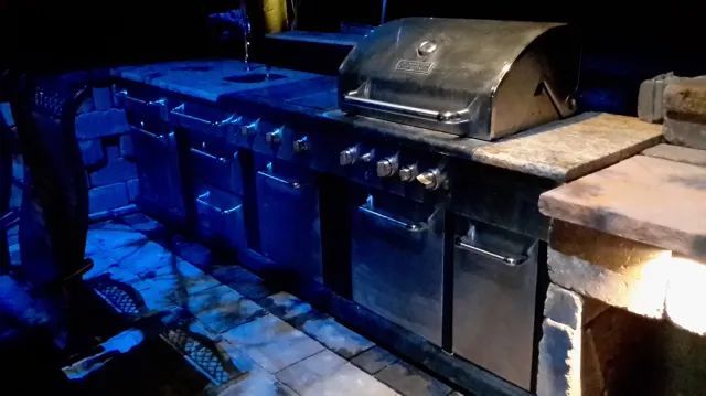 Outdoor kitchen with stainless steel grill, cabinets, and brick counter in dark setting.