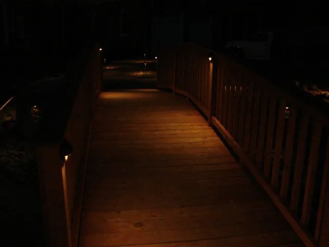 Wooden bridge at night, lit by small lights along the railings and pathway, casting warm glow.
