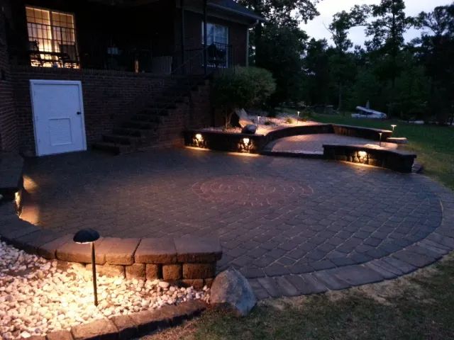 Brick patio with built-in seating, illuminated by landscape lighting at night, adjacent to a house.