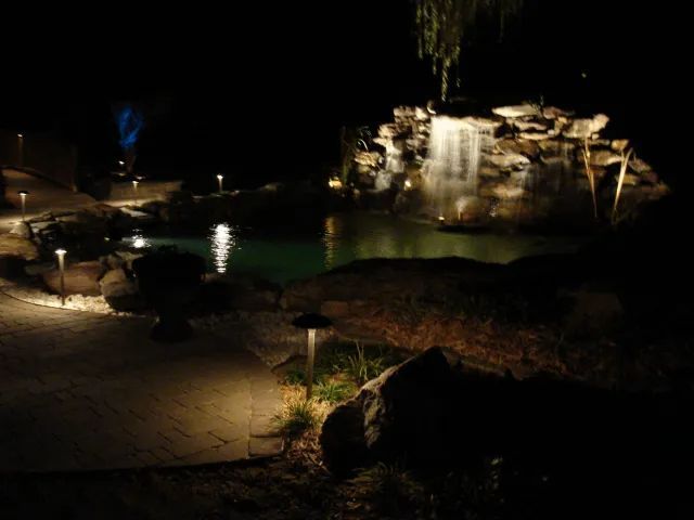 Night view of a lit-up pond with a waterfall, pathway, and landscape lights. Dark setting.