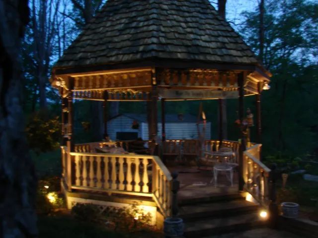 Lit gazebo at dusk, wooden with ornate details, steps leading up.