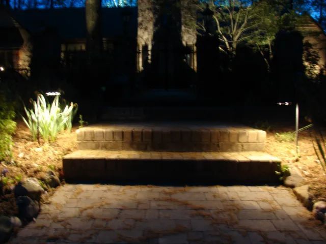 Brick pathway and steps leading to a house, illuminated by outdoor lights at night.