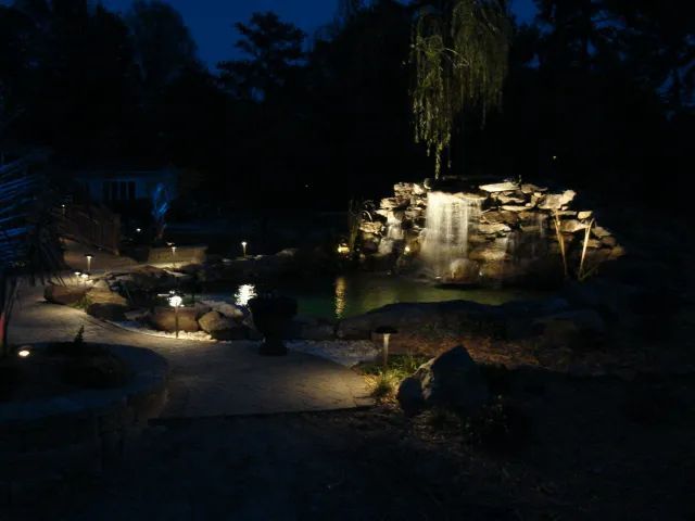 Nighttime outdoor scene with illuminated waterfall and pond surrounded by rocks and lights.