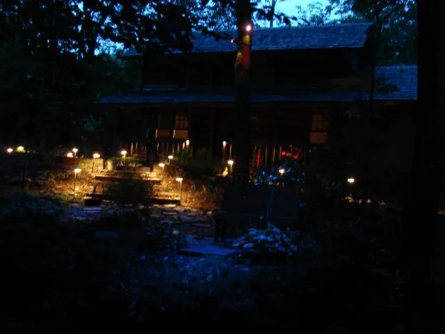 A dark, tree-covered house at night, lit by small pathway lights.
