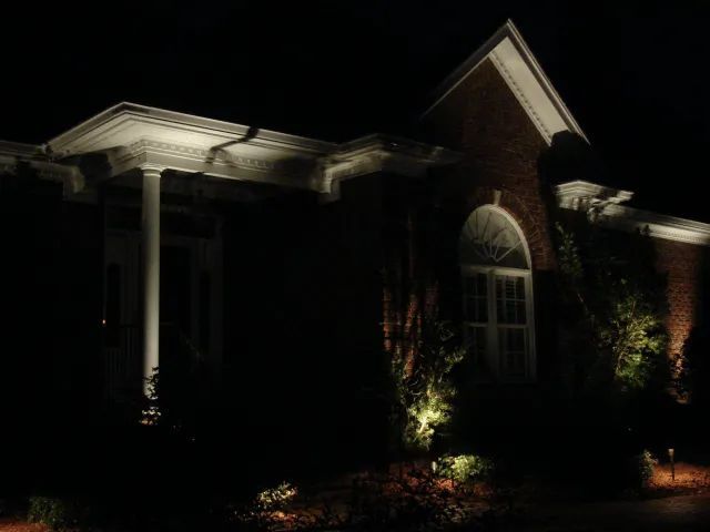 House exterior at night, lit by spotlights. Brick, white trim, and plants are illuminated.