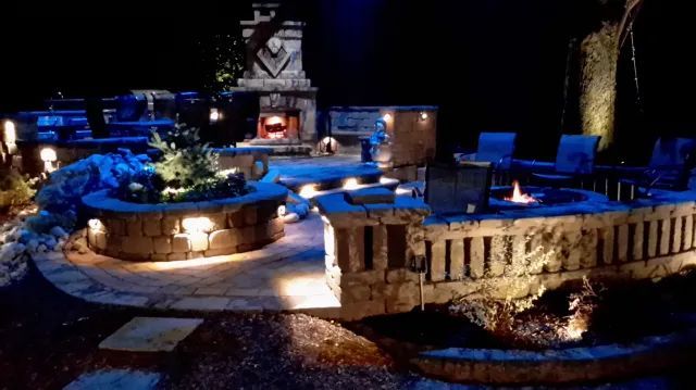 Nighttime outdoor stone patio with built-in seating, fire pits, and fireplace, illuminated by soft lighting.
