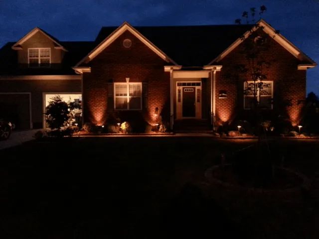 House at dusk, illuminated by outdoor lights; brown brick, white trim, dark blue sky.