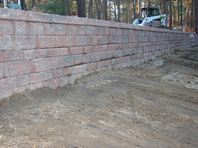 A long, reddish-brown retaining wall with a flat top, dirt in front. A small tractor is in the background.