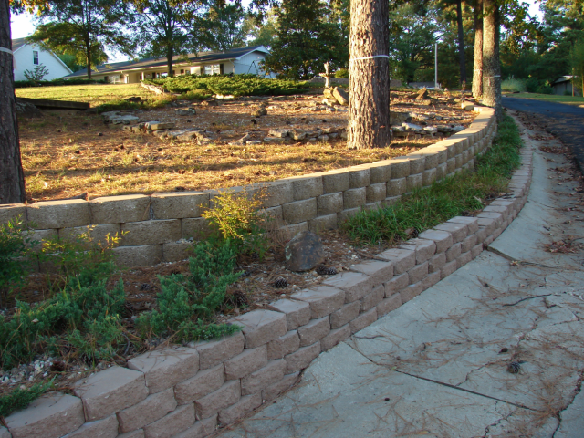 A retaining wall of brick-like blocks borders a driveway next to a grassy area with trees.