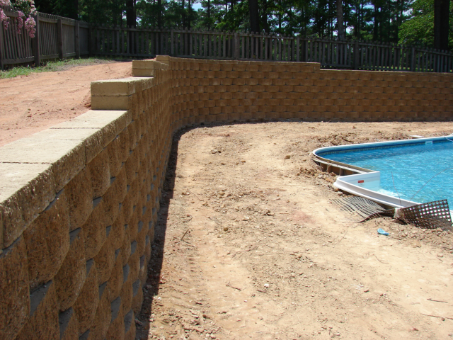 Retaining walls made of tan blocks surround a dirt area and a swimming pool with blue water.