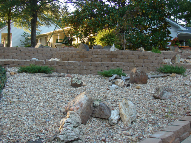 Stone landscaping with retaining walls, rocks, gravel, and small plants in front of a house.