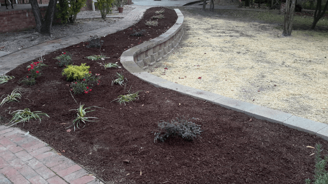 A brick pathway curves past a landscaped area with mulch, plants, and a concrete border.