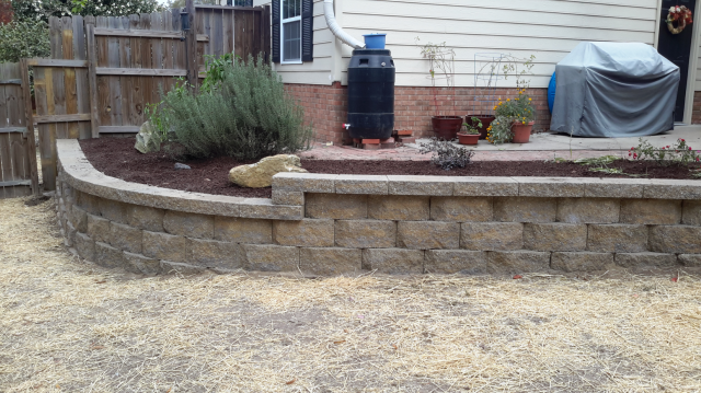Stone retaining wall bordering a patio with plants and mulch, beside a house and fence.