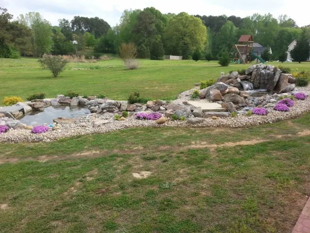 Pond and waterfall feature in a grassy yard, surrounded by rocks and purple flowers. Trees in the background.