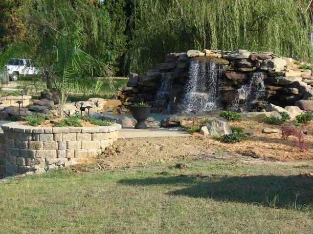 Waterfall feature with rocky facade, flowing into a pond area; brick-lined planter and greenery in a yard.