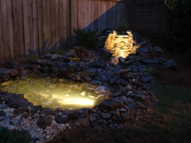 Night view of a lit backyard pond with waterfall, surrounded by rocks and a wooden fence.