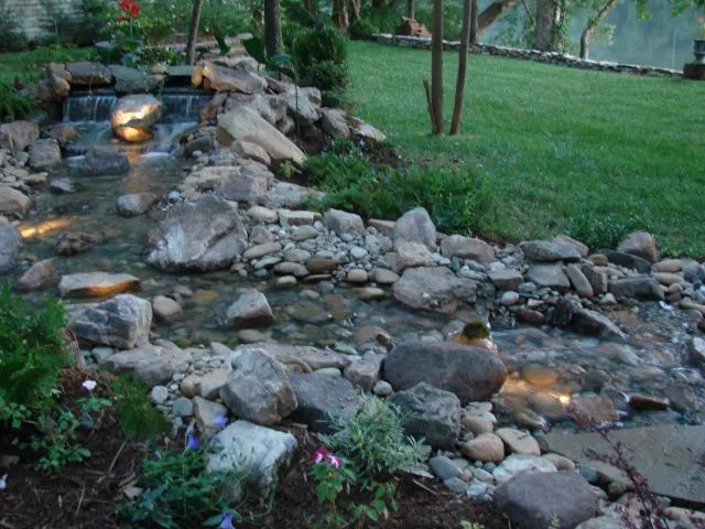 A rocky water stream cascading into a shallow pool, illuminated by soft lights, next to green grass.