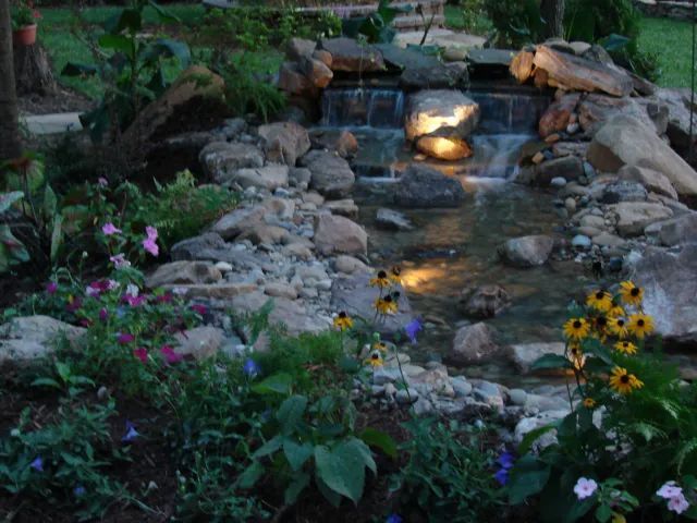 Stone waterfall feature with illuminated water, surrounded by rocks and flowers.