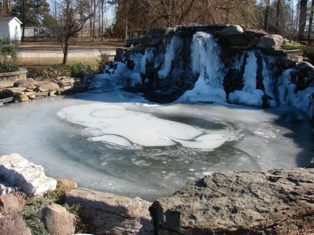 Frozen waterfall cascading into a partially iced pond surrounded by rocks and a small building in the background.