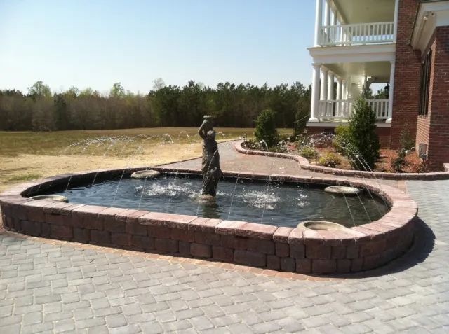 Fountain with statue in a brick-lined pond, in front of a house, with water spraying.