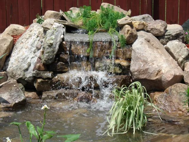 Small backyard waterfall cascading over rocks into a pond, surrounded by greenery.
