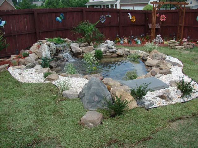 Backyard pond with waterfall, surrounded by rocks, white stones, and green grass.