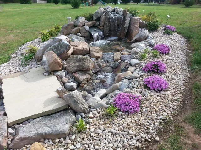 Rock waterfall and pond in a backyard, surrounded by rocks, pebbles, and purple flowering plants.