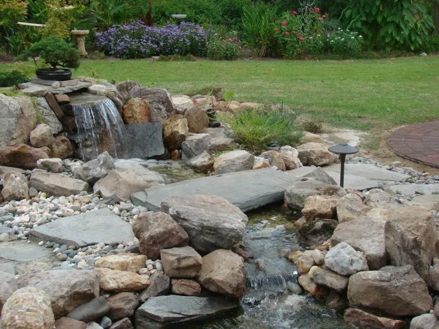Waterfall feature in a landscaped garden, with rocks, bridge, and flowing water.