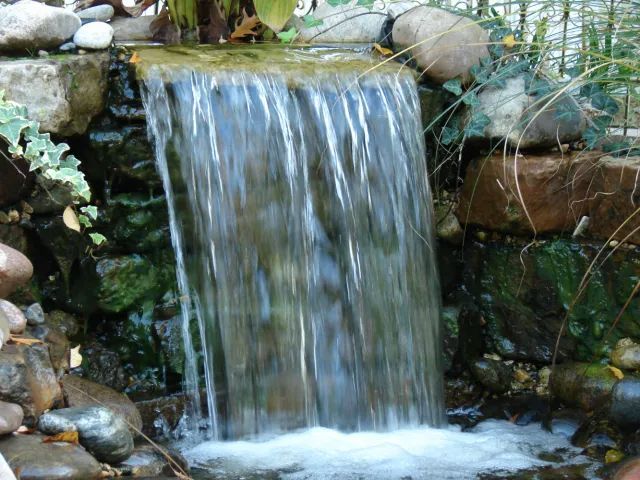 Small stone waterfall flowing into a pool, surrounded by rocks and greenery.