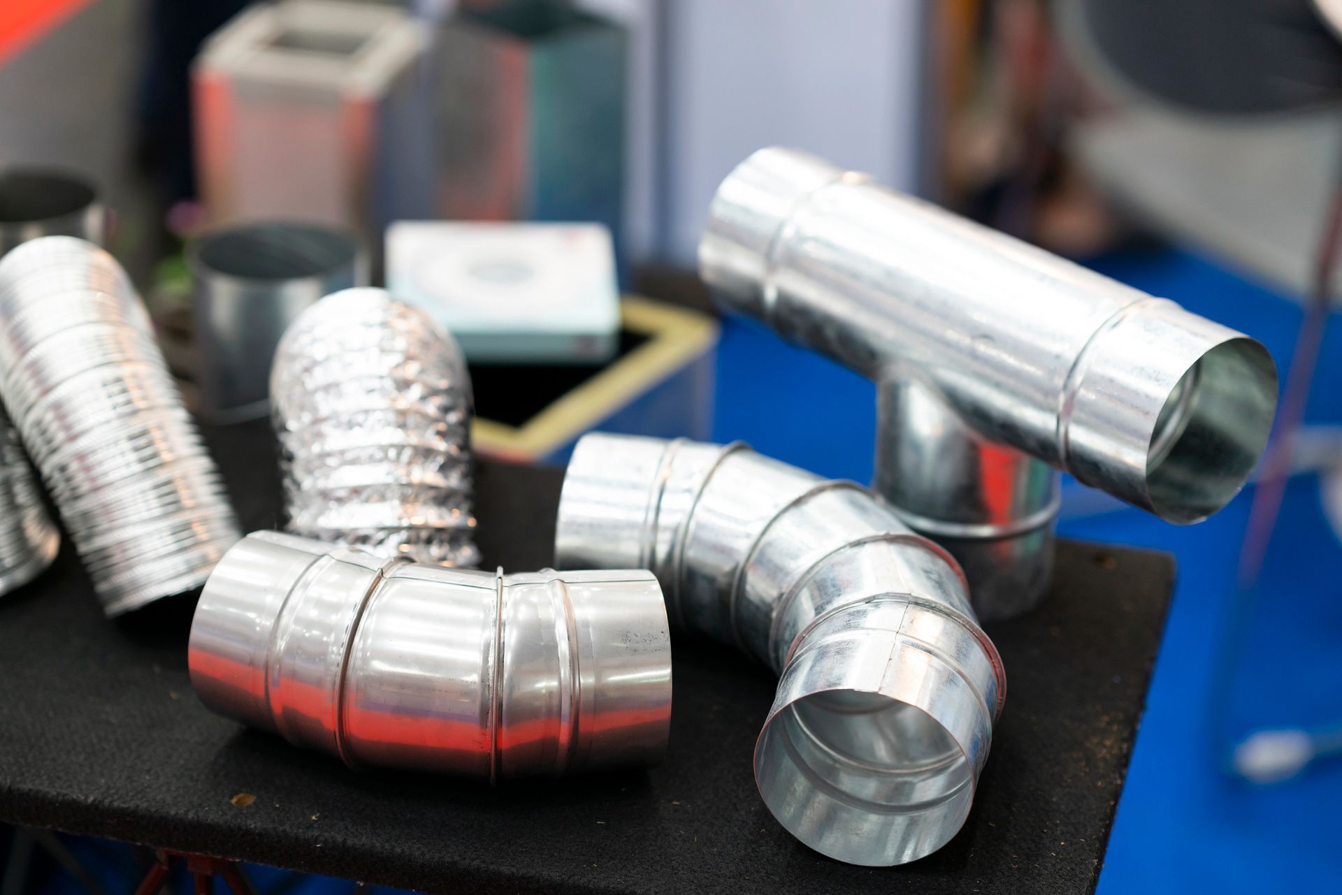 Componenti di condotti metallici esposti su una superficie nera; diverse forme, tra cui gomiti, sezioni dritte e tubi flessibili.