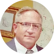 Man with glasses, wearing a suit, smiling slightly, in front of a painting.