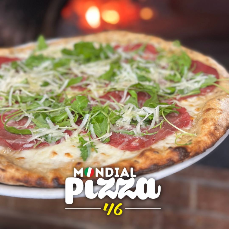 Una pizza con prosciutto e rucola su un piatto bianco, con un forno acceso sullo sfondo, intitolata Mondial Pizza 46.