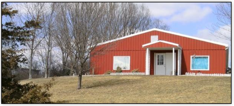 A red metal-sided building with a small entrance porch sits on a grassy hill under a bright, cloudy sky.