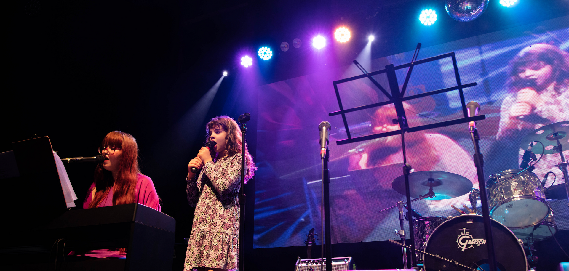 Two women are singing on a stage in front of a large screen.