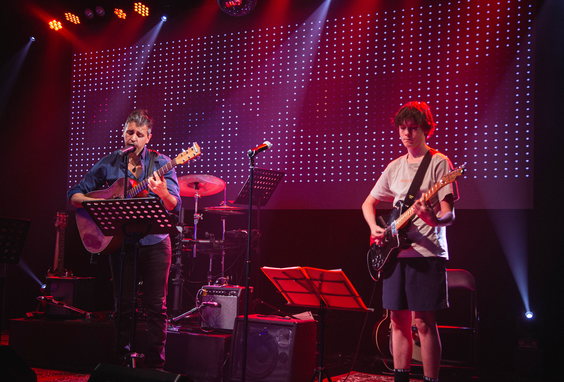 Two men are playing guitars on a stage with purple lights behind them