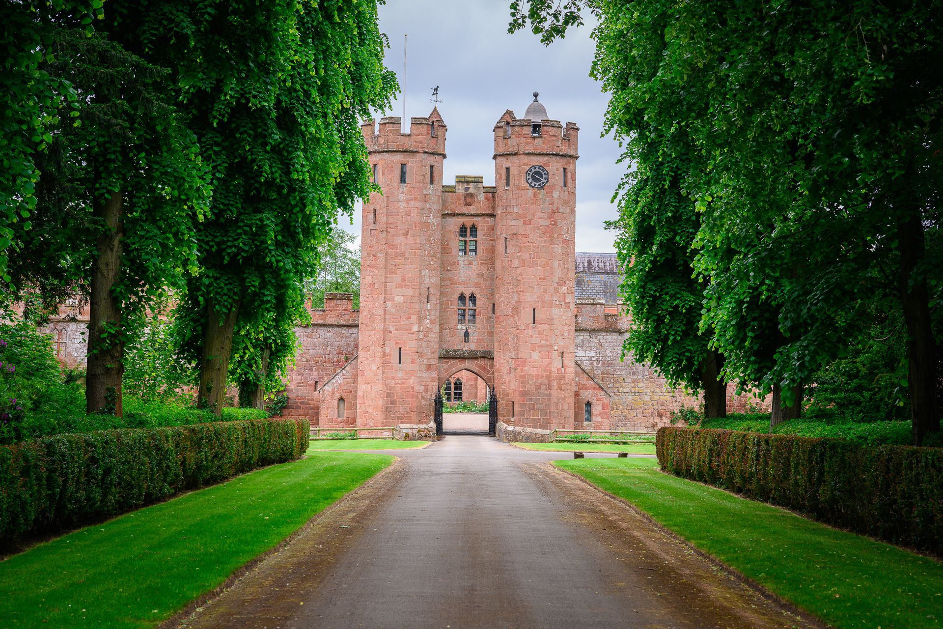 A tree-lined gravel path leads to a historic red-brick gatehouse with two round towers under a cloudy sky.