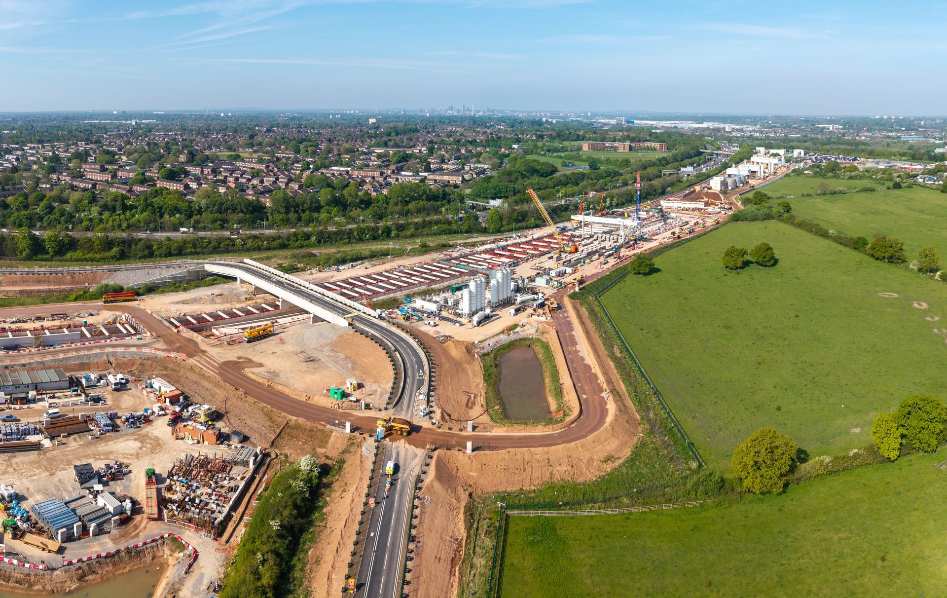 Aerial view of a construction site with machinery, a bridge, and green fields. Blue sky in the background.